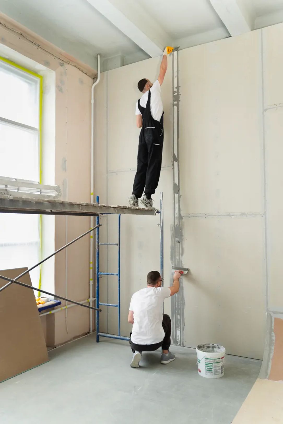 Two drywall installers finishing a wall, one on scaffolding and one applying mud below