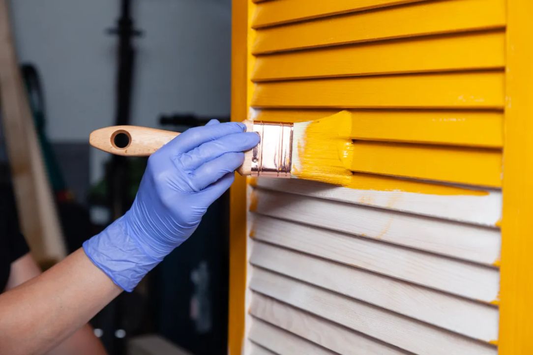 Painter's gloved hand brushing yellow paint onto louvered wood